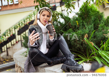 stylish adult woman in headphones drinks coffee and looks at the screen of a mobile phone while 94876008