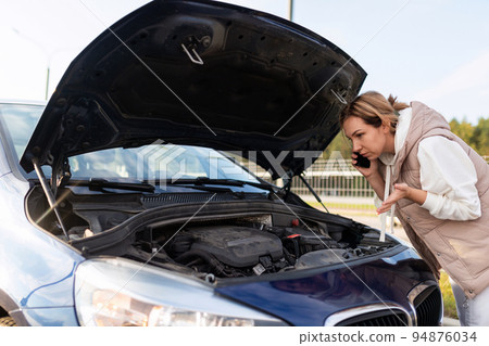 confused female driver looking under the hood talking on a mobile phone and trying to understand the 94876034