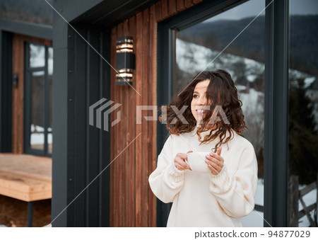 Curly young woman resting on terrace of modern barn house in the mountains. Happy female tourist holding cup of tea, enjoying in new cottage in winter. 94877029