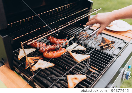 Pieces of white bread and sausages are grilling on the open fire outside. Hand of young woman grilling food, gas grill 94877445