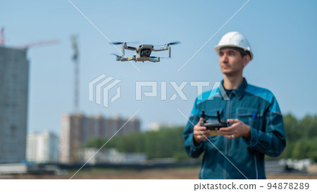 A man in a helmet and overalls controls a drone at a construction site. The builder carries out technical oversight. 94878289