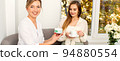 Beautician offering product for the young woman holding a white plastic jar with a cream sitting on the sofa 94880554
