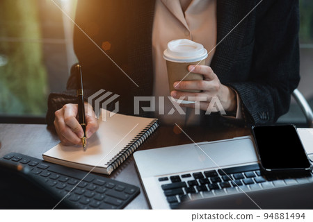 Business woman using laptop to work and writing on notebook with pen in office with mobile next to laptop. 94881494
