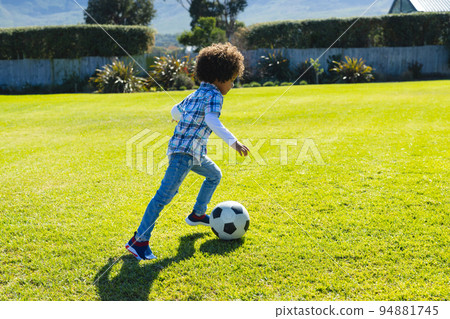 Side view of biracial boy with afro hair running and kicking soccer ball on grassy field in yard Side view of biracial boy with afro hair running and kicking soccer ball on grassy field in yard 94881745
