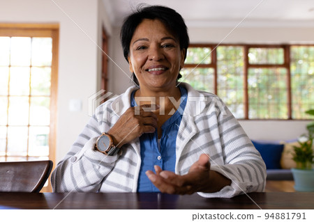 Smiling biracial mature woman with short hair pointing finger on neck while sitting at table Smiling biracial mature woman with short hair pointing finger on neck while sitting at table 94881791