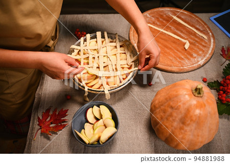 Top view of a housewife in beige chef's apron, decorating the classic American homemade festive pie with a crunchy pastry lattice. Thanksgiving Day. Making tasty pumpkin apple pie with flaky crust. Top view of a housewife in beige chef's apron, decorating the classic American homemade festive pie with a crunchy pastry lattice. Thanksgiving Day. Making tasty pumpkin apple pie with flaky crust. 94881988