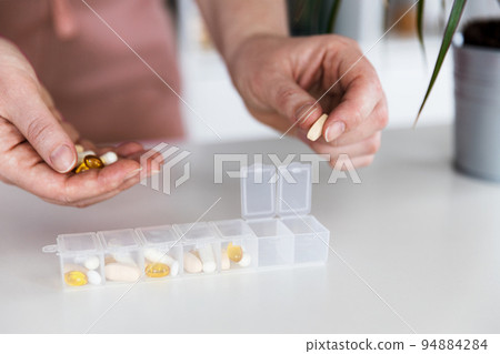 Female hands sorting pills. Young woman getting her daily vitamins at home. Closeup of medical pill box with doses of tablets for daily take a medicine with different white, yellow drugs and capsules Female hands sorting pills. Young woman getting her daily vitamins at home. Closeup of medical pill box with doses of tablets for daily take a medicine with different white, yellow drugs and capsules 94884284