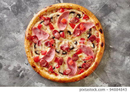 Freshly baked pizza with hunting sausages, ham, mushrooms and tomatoes on gray stone surface 94884843