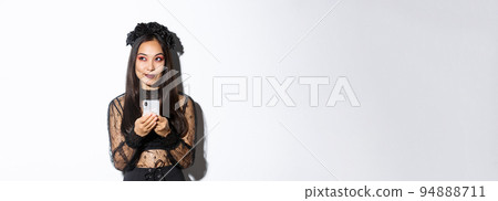 Thoughtful pretty asian girl in gothic elegant dress holding smartphone while thinking, looking at upper left corner, standing over white background Thoughtful pretty asian girl in gothic elegant dress holding smartphone while thinking, looking at upper left corner, standing over white background 94888711