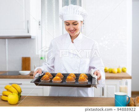Happy female chef in white uniform standing with tray of muffins in private kitchen 94888922