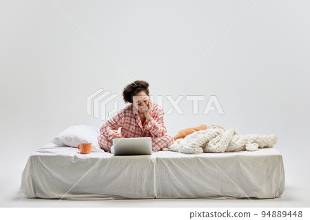 Portrait of young charming girl in home wear sitting on bed at home isolated on gray background. Ideas, inspiration, imagination. Art, beauty, youth 94889448
