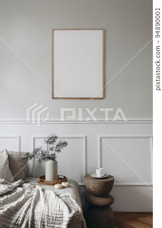 Moody Christmas interior. Blank vertical wooden picture frame mockup. Moulding wall background.. Bed, sofa with linen cushions, blanket. Pine tree branches in vase. Wooden side table, cup of coffee. 94890001