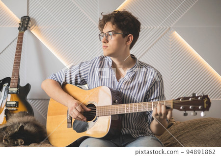Modern man playing acoustic guitar learning musical performance sitting on couch at home Modern man playing acoustic guitar learning musical performance sitting on couch at home 94891862