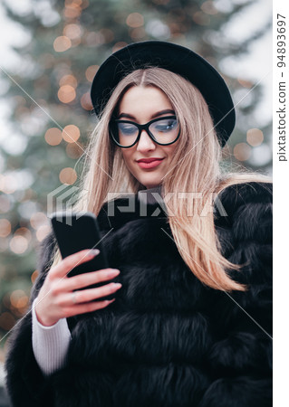 Beautiful girl in glasses and a hat uses a smartphone on a background of Christmas tree lights 94893697