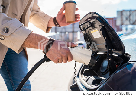 Stylish man inserts plug of the charger into the socket of electric car close-up 94893993
