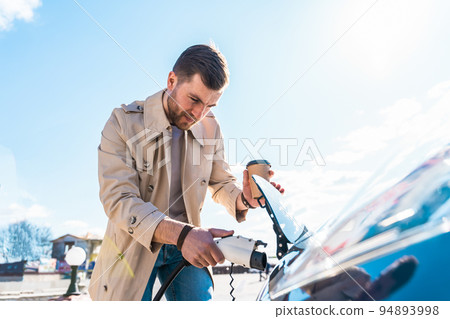 Stylish man with coffe cup in hand inserts plug into the electric car charging socket 94893998