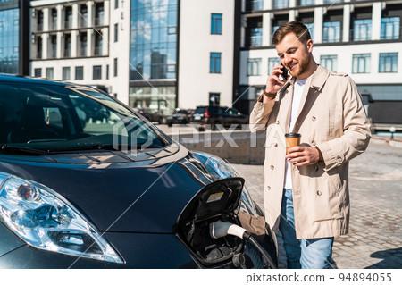 Handsome man talks on the phone and drinks coffee while his car is charging at the station 94894055