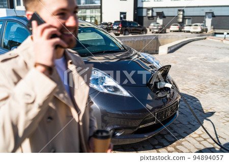 Handsome man talks on the phone and drinks coffee while his car is charging at the station Handsome man talks on the phone and drinks coffee while his car is charging at the station 94894057