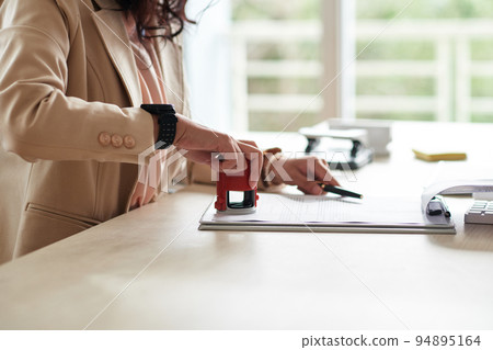 Close-up of young businesswoman putting stamp on document at table at office Close-up of young businesswoman putting stamp on document at table at office 94895164