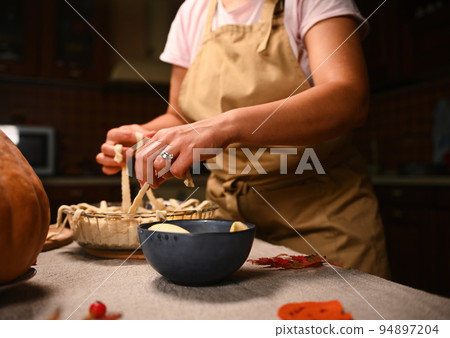Cropped view of pastry chef in beige apron standing at table with linen tablecloth, decorating homemade pie for Thanksgiving dinner, holding pastry strips, making lattice pattern on top of pumpkin pie Cropped view of pastry chef in beige apron standing at table with linen tablecloth, decorating homemade pie for Thanksgiving dinner, holding pastry strips, making lattice pattern on top of pumpkin pie 94897204