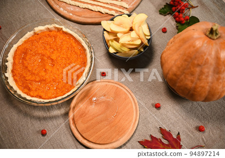 Top view festive Thanksgiving pie, pastry stripes on wooden board, slices of apple in bowl, autumn maple leaves, viburnum berries, whole ripe pumpkin on a linen tablecloth. Rustic still life with food 94897214