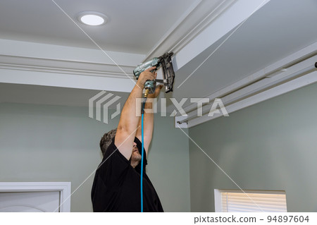 The a professional carpenter nailing wooden crown molding between the walls and the ceiling of a newly constructed house The a professional carpenter nailing wooden crown molding between the walls and the ceiling of a newly constructed house 94897604