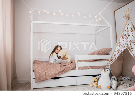 Cute little girl in pajama hugging her toy bear on the scandinavian wooden house frame bed at home Cute little girl in pajama hugging her toy bear on the scandinavian wooden house frame bed at home 94897816