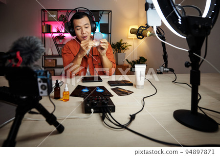 Young adult Asian man wearing headphones sitting desk in home studio demonstrating new face powder while doing online streaming 94897871