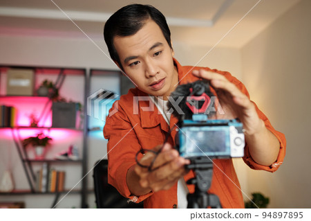 Horizontal selective focus medium portrait of Asian male influencer preparing for video recording adjusting camera on tripod Horizontal selective focus medium portrait of Asian male influencer preparing for video recording adjusting camera on tripod 94897895