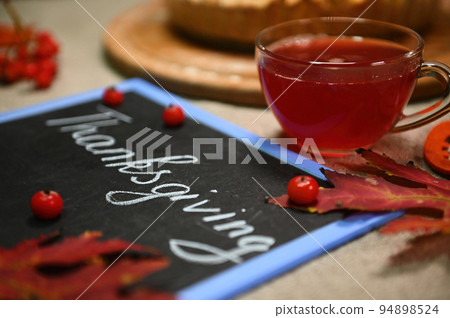 Selective focus. Still life with a glass teacup with healthy antioxidant hot herbal drink, blurred foreground with dry fallen autumn maple leaves and a blackboard with lettering Thanksgiving Day. Selective focus. Still life with a glass teacup with healthy antioxidant hot herbal drink, blurred foreground with dry fallen autumn maple leaves and a blackboard with lettering Thanksgiving Day. 94898524