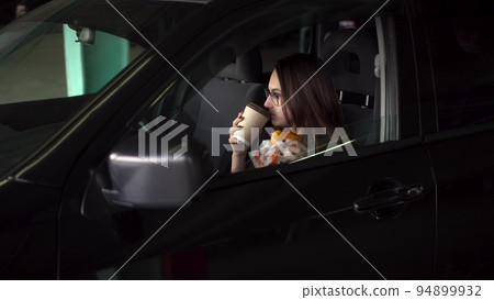 Businesswoman has lunch in the car with a quick meal. A respectable woman in glasses and a suit eats a cheeseburger and drinks coffee. 94899932