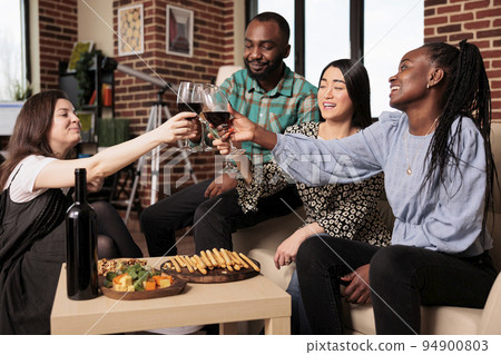 Culturally diverse group of adults, men, women toasting, seated at living room couches, drinking glasses of wine. Ethnically different people bunch enjoying alcohol, eating bread sticks. 94900803