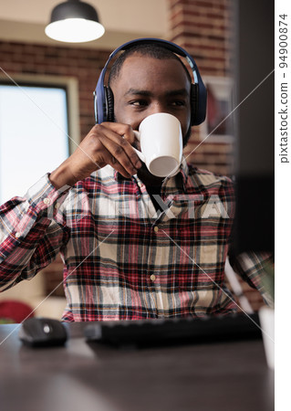 Young adult working from home on computer and listen to music or podcast show online on headphones. Man using headset and browsing internet website, doing remote freelance job. 94900874