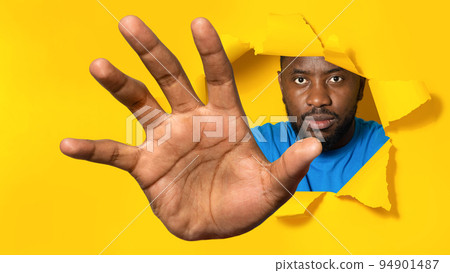 Serious african american man reaching through hole in torn yellow paper, trying to grab something Serious african american man reaching through hole in torn yellow paper, trying to grab something 94901487