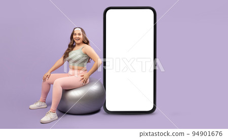 Happy european young lady plus size do exercises on fitness ball near big smartphone with blank screen 94901676