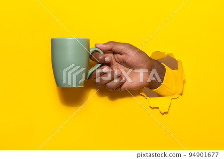 African american man holding cup of hot beverage through torn yellow paper background, closeup, copy space 94901677