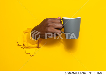 Black male hand holding a mug of coffee through torn yellow paper background, closeup, copy space 94901689