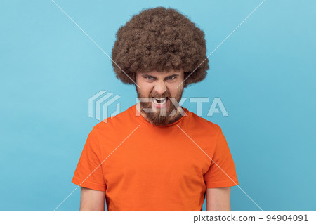Portrait of angry man with Afro hairstyle wearing orange T-shirt looking at camera, having aggressive facial expression, expressing negative, hatred. Indoor studio shot isolated on blue background. 94904091