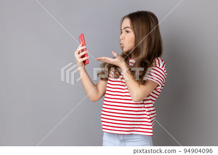 Portrait of pretty little girl wearing striped T-shirt sending kiss on smartphone camera while communicating by video call or streaming vlog. Indoor studio shot isolated on gray background. 94904096