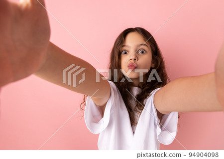 Portrait of adorable little girl wearing white T-shirt sending air kisses while taking selfie POV, flirting, expressing romantic feelings. Indoor studio shot isolated on pink background. Portrait of adorable little girl wearing white T-shirt sending air kisses while taking selfie POV, flirting, expressing romantic feelings. Indoor studio shot isolated on pink background. 94904098