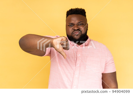 Portrait of sad upset bearded man wearing pink shirt showing thumb down, expressing bad negative emotions, dislike, frowning face. Indoor studio shot isolated on yellow background. Portrait of sad upset bearded man wearing pink shirt showing thumb down, expressing bad negative emotions, dislike, frowning face. Indoor studio shot isolated on yellow background. 94904099
