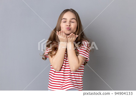Portrait of adorable beautiful little girl wearing striped T-shirt sending air kiss with pout lips, experiencing first romantic feelings. Indoor studio shot isolated on gray background. 94904133