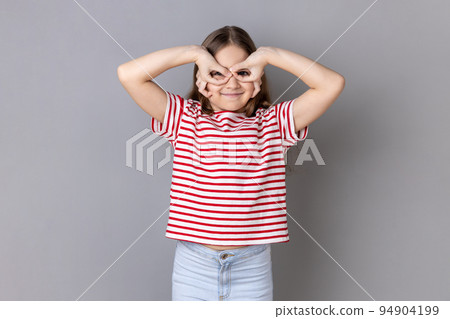 Portrait of funny positive little girl wearing striped T-shirt making binoculars gesture, looking far away with cute smile. Indoor studio shot isolated on gray background. 94904199