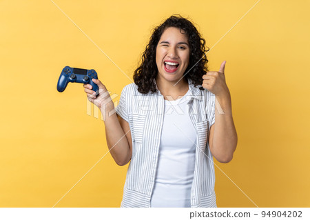 Portrait of joyful beautiful woman with dark wavy hair holding joystick in hands and showing thumb up, likes playing games on joypad. Indoor studio shot isolated on yellow background. 94904202