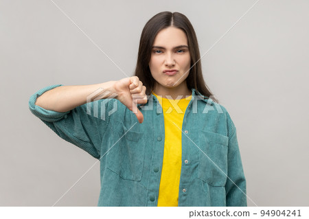Dissatisfied woman showing thumb down, disapproval sign, gesturing dislike to bad service, disagree with suggestion, wearing casual style jacket. Indoor studio shot isolated on gray background. 94904241