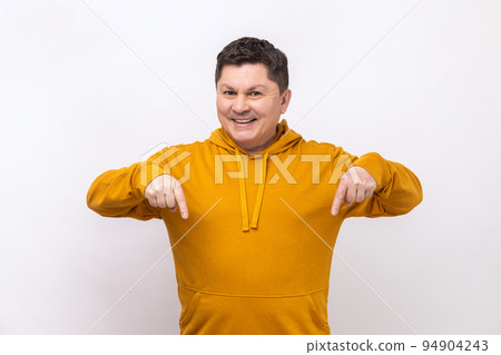 Cheerful delighted man pointing fingers down, paying attention to advertising area, blogger recommending to subscribe, wearing urban style hoodie. Indoor studio shot isolated on white background. 94904243