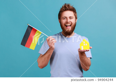 Portrait of positive optimistic handsome bearded man holding toy paper house and flag of germany in hand, moving into another country. Indoor studio shot isolated on blue background. 94904244
