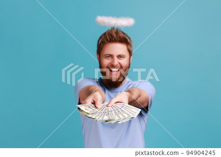Portrait of rich handsome satisfied bearded angelic man with holy nimbus over his head holding out big fan of dollar banknotes, win the lottery. Indoor studio shot isolated on blue background. 94904245