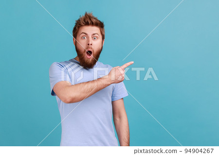 Portrait of surprised shocked bearded man pointing aside, showing blank copy space for idea presentation, commercial text, keeps mouth open. Indoor studio shot isolated on blue background. 94904267