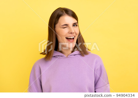 Portrait of positive optimistic woman standing and winking playfully, positive expression, looking at camera, wearing purple hoodie. Indoor studio shot isolated on yellow background. 94904334